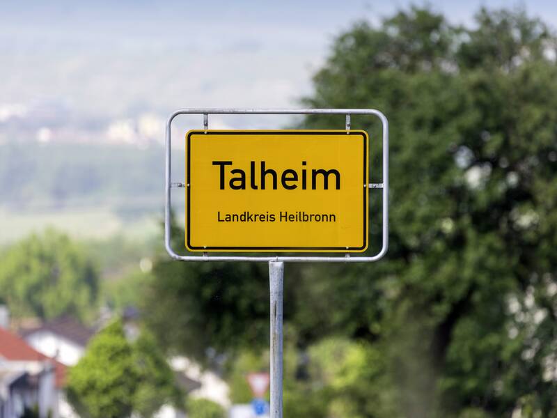 Ein Schild zeigt den Ortsnamen "Talheim, Landkreis Heilbronn" vor einer unscharf wirkenden grünen Landschaft und einem Baum im Hintergrund.