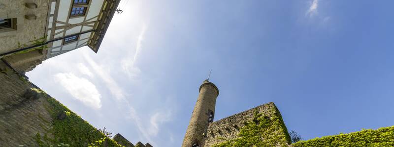 Der Blick von einem Innenhof aus zeigt ein Fachwerkhaus und einen Turm einer alten Burg mit bewachsenen Steinmauern unter einem klaren blauen Himmel.