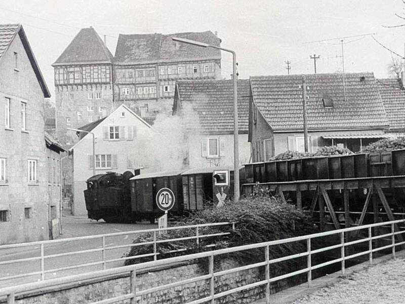 Eine alte Dampflokomotive fährt durch eine von Häusern umgebene Straße, während neben ihr Waggons über eine Brücke verlaufen.