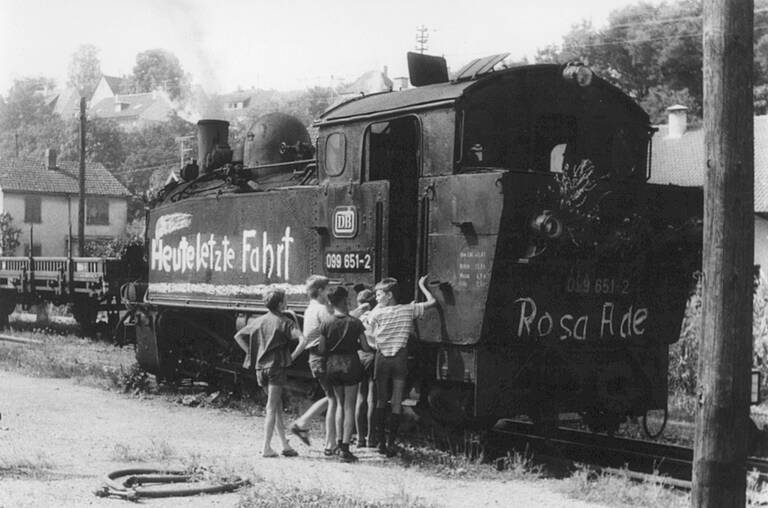 Einige Kinder stehen neben einer Dampflokomotive mit der Aufschrift "Heute letzte Fahrt" und "Rosa Ade".