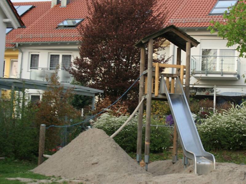 Ein Spielplatz mit einer Rutsche aus Metall neben einem Sandhaufen, im Hintergrund sind Häuser mit roten Dächern zu sehen.