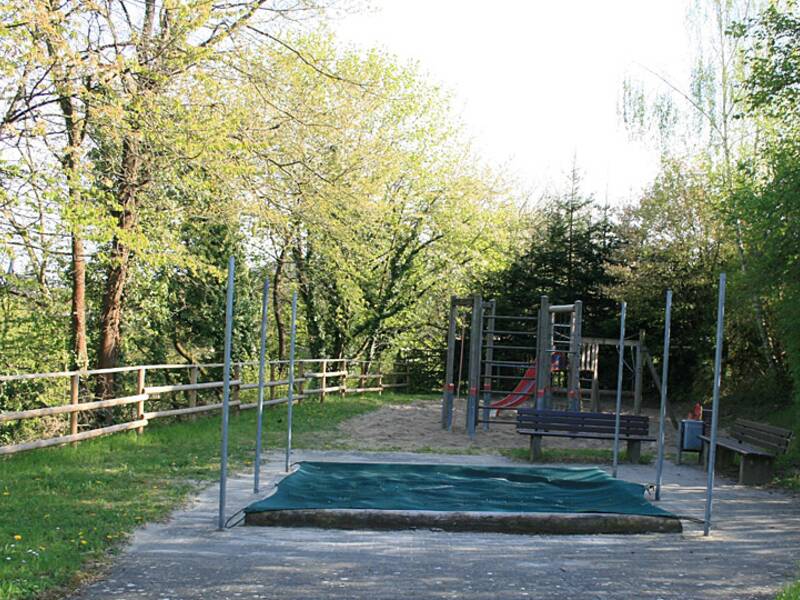 Ein Spielplatz mit Klettergerüst und Rutsche, umgeben von Bäumen, mit einer grünen Matte im Vordergrund.
