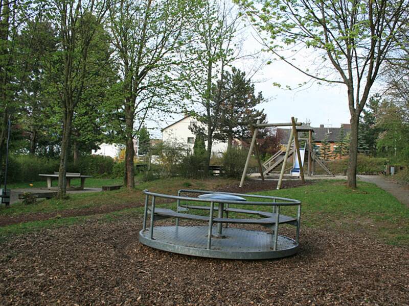 Ein ruhiger Spielplatz mit einem Metallkarussell und einer Schaukel, umgeben von Bäumen und Grünflächen.