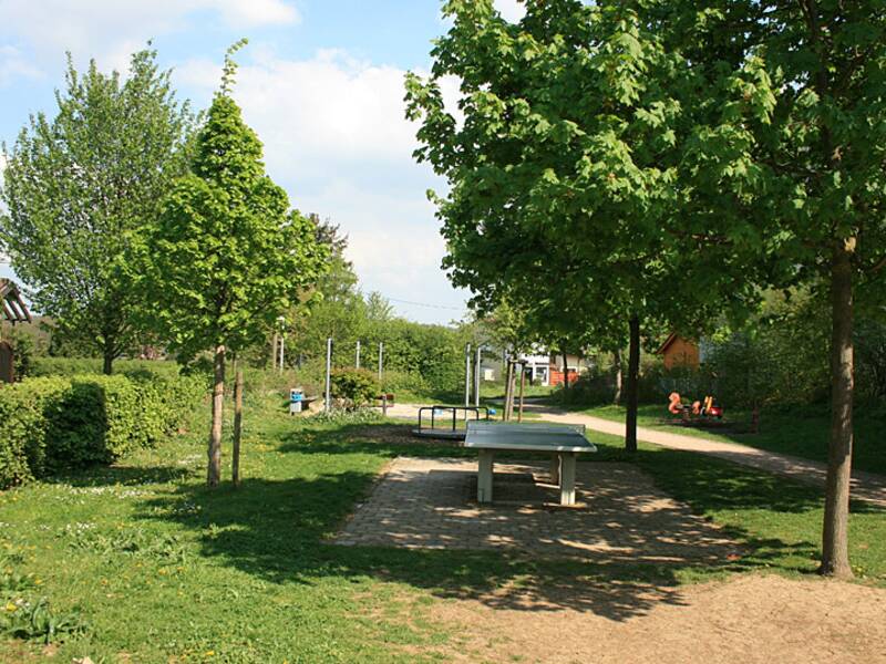 Ein kleiner Park mit einer Tischtennisplatte im Vordergrund, umgeben von grünen Bäumen und einem Spielplatz im Hintergrund.