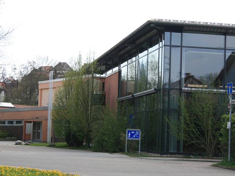 Ein modernes Gebäude mit großen Glasfronten und Bäumen davor steht an einer ruhigen Straße mit einem Gehweg und einem Straßenschild.