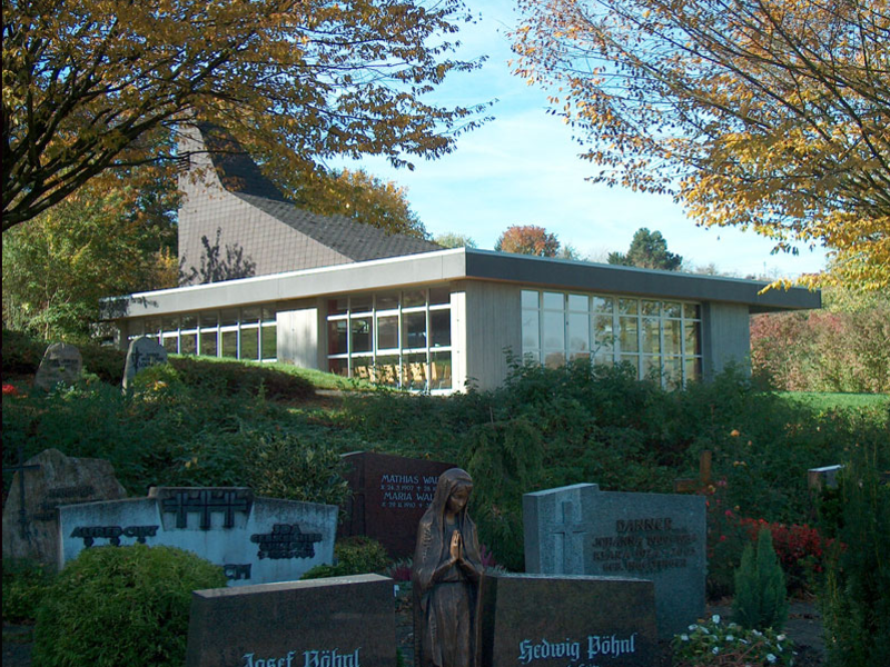 Eine moderne Kapelle mit großen Fenstern steht hinter mehreren Grabsteinen in einer grünen und herbstlichen Umgebung.