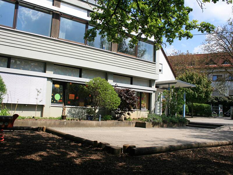 Ein moderner Gebäudekomplex mit großen Fenstern und bepflanzter Umgebung, umgeben von Bäumen, unter einem blauen Himmel.