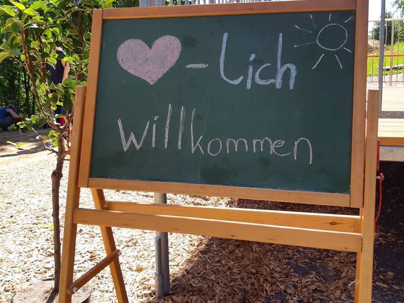 Eine grüne Tafel auf einem Holzständer steht draußen und zeigt in Kreideschrift "Herzlich willkommen" mit einem Herzsymbol und einer Sonne.