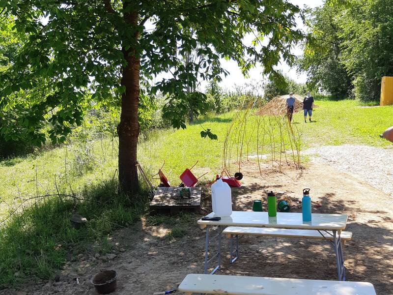 Ein sonniger Garten mit einem Baum im Vordergrund, einem Tisch und Bänken sowie zwei Personen, die in der Ferne neben einem Weidenbau laufen.