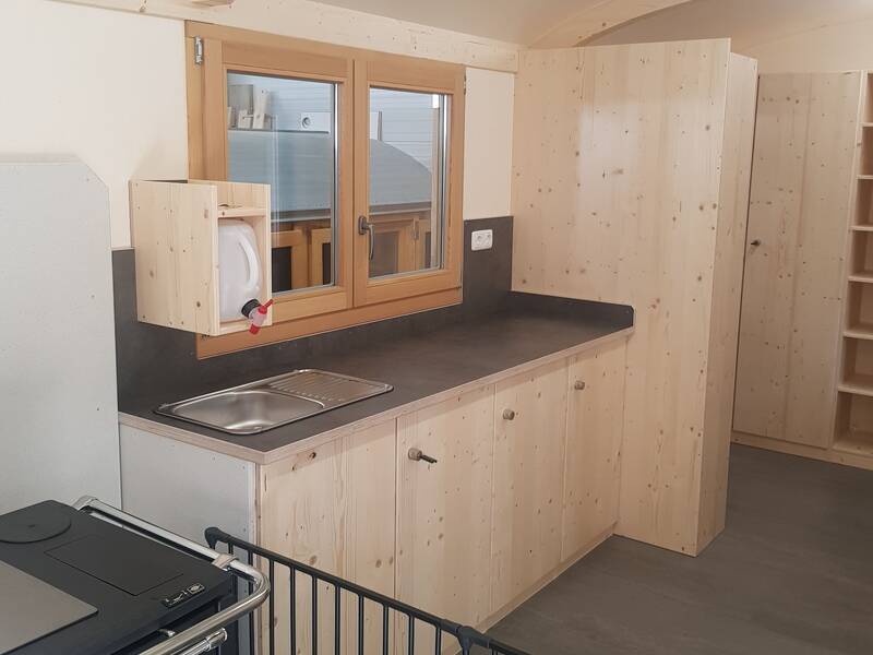 Eine Holzküche mit einem Waschbecken, Fenster, Schränken und einem Holzofen im Vordergrund.