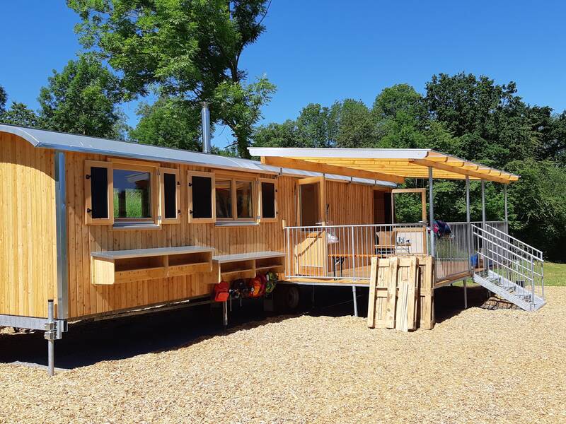 Ein modern gestalteter, hölzerner Bauwagen steht auf einem mit Holzschnitzeln bedeckten Platz in einer grünen Umgebung.