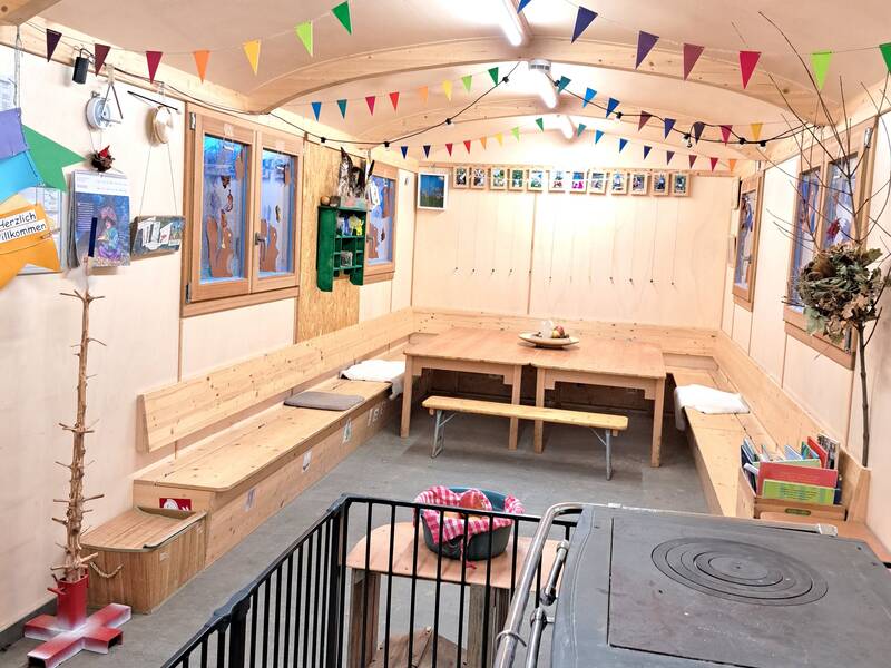 Ein gemütlicher Raum mit Holzbänken, einem Tisch, bunten Wimpelgirlanden an der Decke und verschiedenen Dekorationen an den Wänden.
