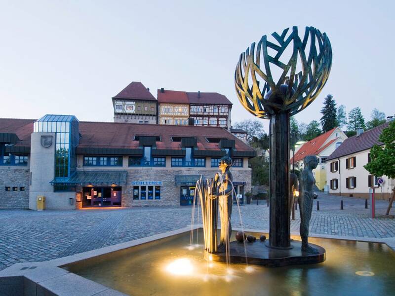 Ein Brunnen mit modernen Skulpturen steht auf einem gepflasterten Platz, umgeben von traditionellen Fachwerkhäusern und modernen Gebäuden.