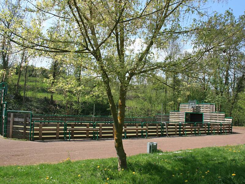 Eine umzäunte, leere Spielfläche mit einem Basketballkorb im Hintergrund, umgeben von Bäumen und Wiese unter einem blauen Himmel.