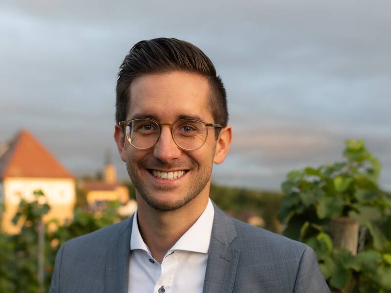 Bürgermeister Pascal Bopp im Anzug steht vor einem Hintergrund mit Weinreben und einem Gebäude mit rotem Dach.